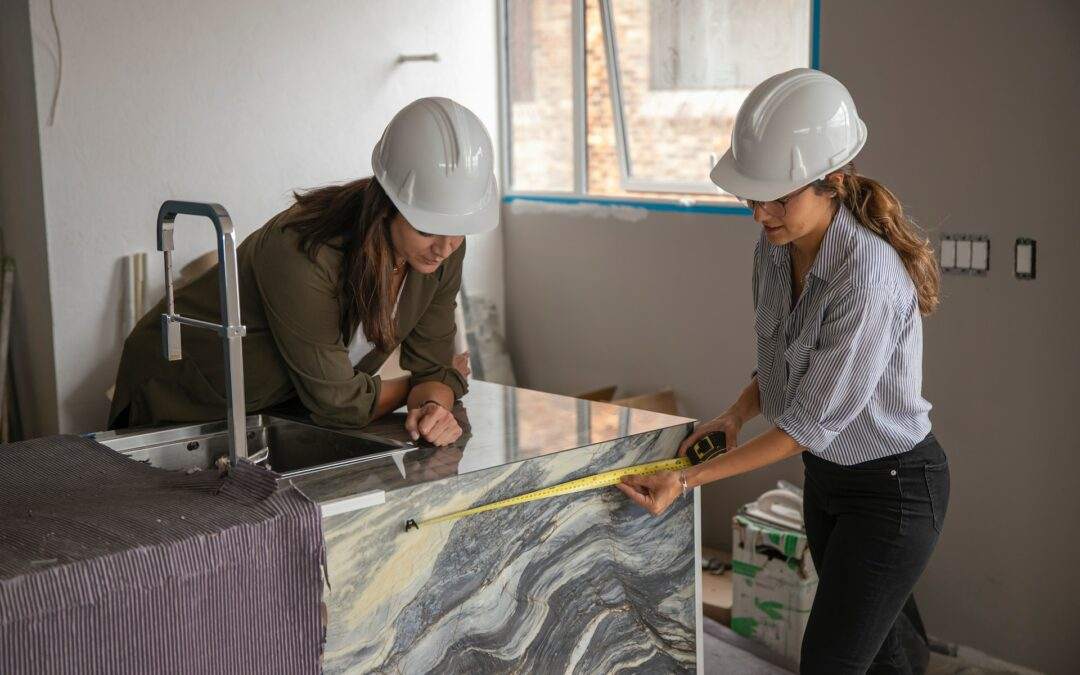 Two women, employed by Riding Homes, a reputable custom home builder in the Southern Utah Valley, are measuring a marble countertop while wearing hard hats.